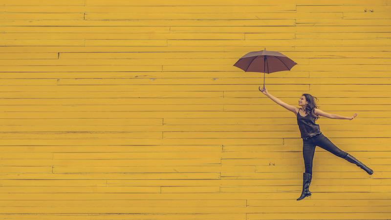 Woman jumps holding an umbrella in front of a yellow wall